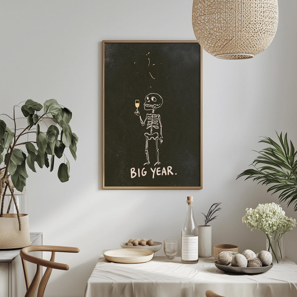 a dining table with a white tablecloth, a bowl of fruit, a bottle, and a vase with flowers. Above the table, there is a framed poster with a skeleton holding a wine glass and the text "BIG YEAR."