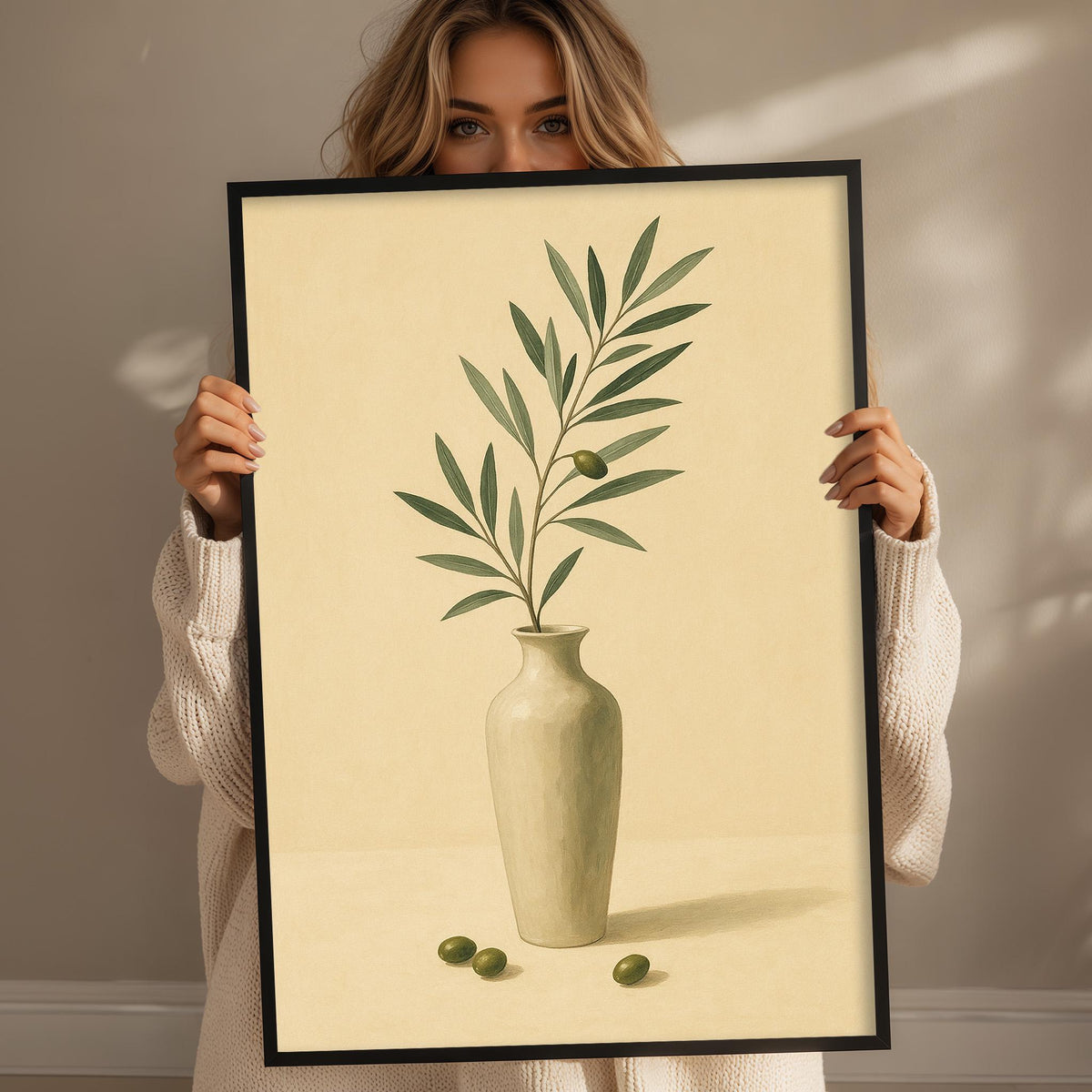 A woman holds up a framed painting of a vase with a plant and three olives on a table.