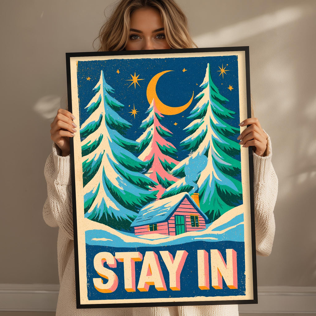 A woman holding a framed poster with a winter scene depicting a cabin and pine trees under a crescent moon and stars.