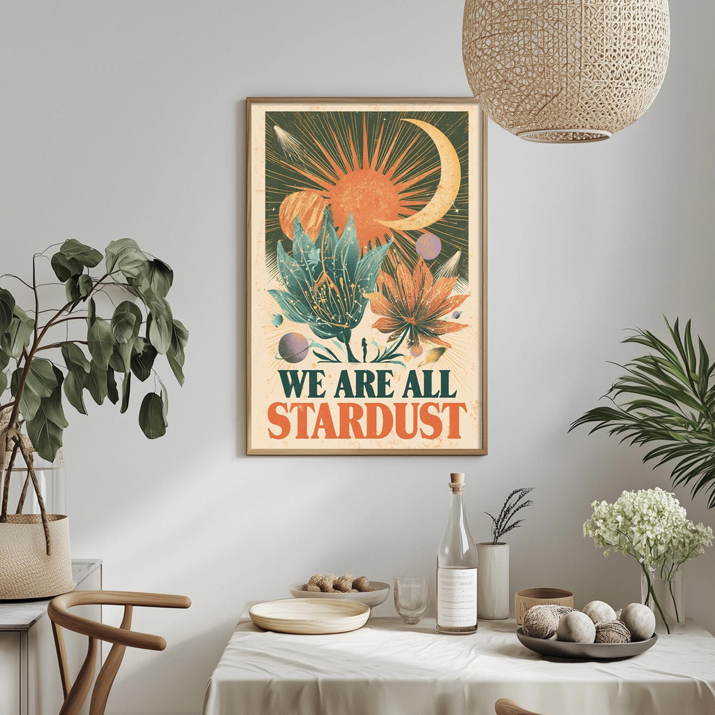 a framed poster with a vibrant, colorful design hanging on a wall above a dining table. The poster displays the text "WE ARE ALL STARDUST" in bold, orange and green lettering. The dining table is set with various items, including a bottle, a bowl, and a vase, creating a cozy and inviting atmosphere.
