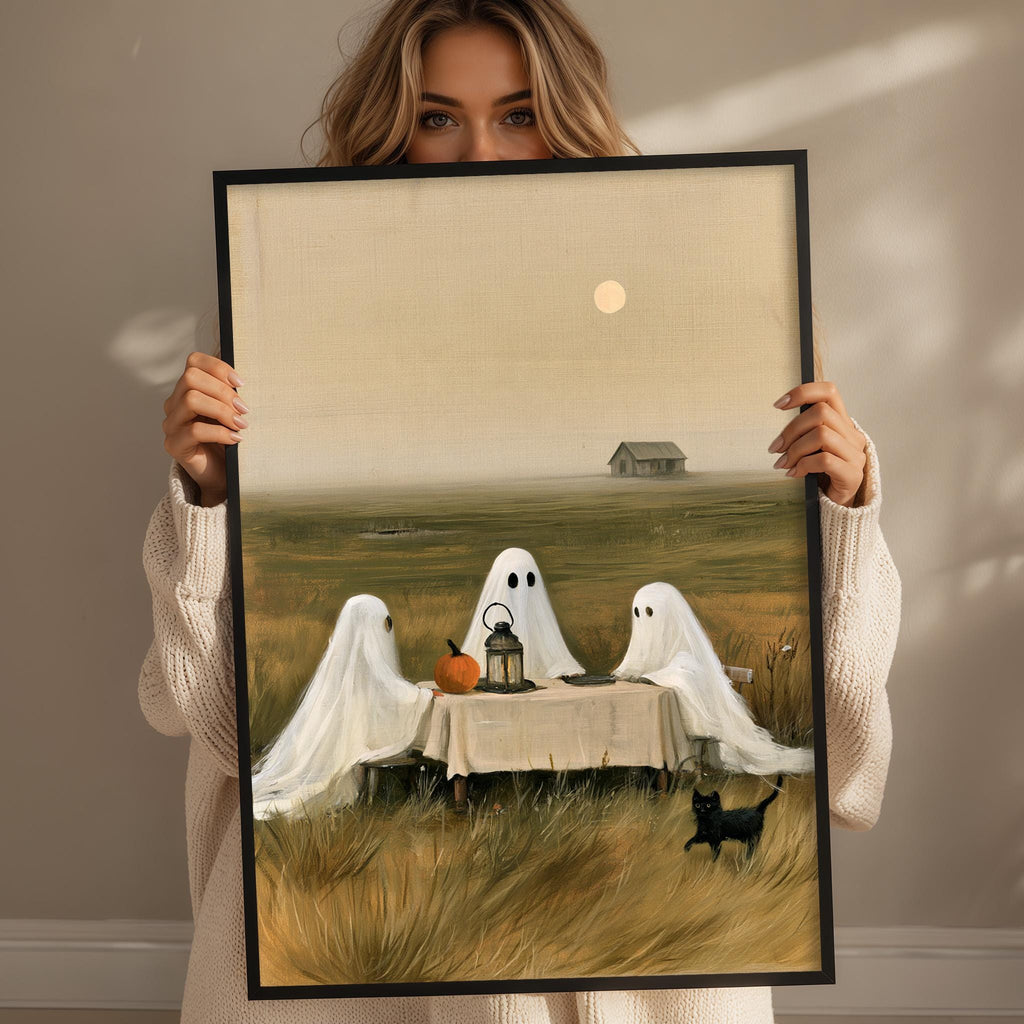 A woman holds up a framed painting depicting three ghostly figures sitting at a table with a pumpkin and a cat, set against a rural landscape with a house and a full moon in the background.