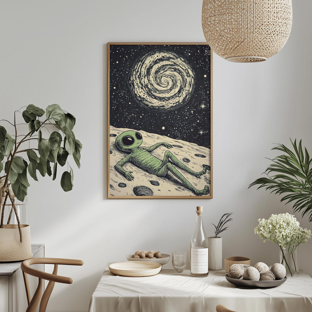 a framed illustration of an alien lying on a moon-like surface, surrounded by a starry night sky. The alien appears to be resting or sleeping, with a spiral galaxy in the background. The scene is set against a white wall, with a potted plant and a vase of flowers adding a touch of nature to the space. The table is set with various items, including a bottle, a bowl, and a plate, suggesting a meal or gathering taking place in this unique and whimsical setting.