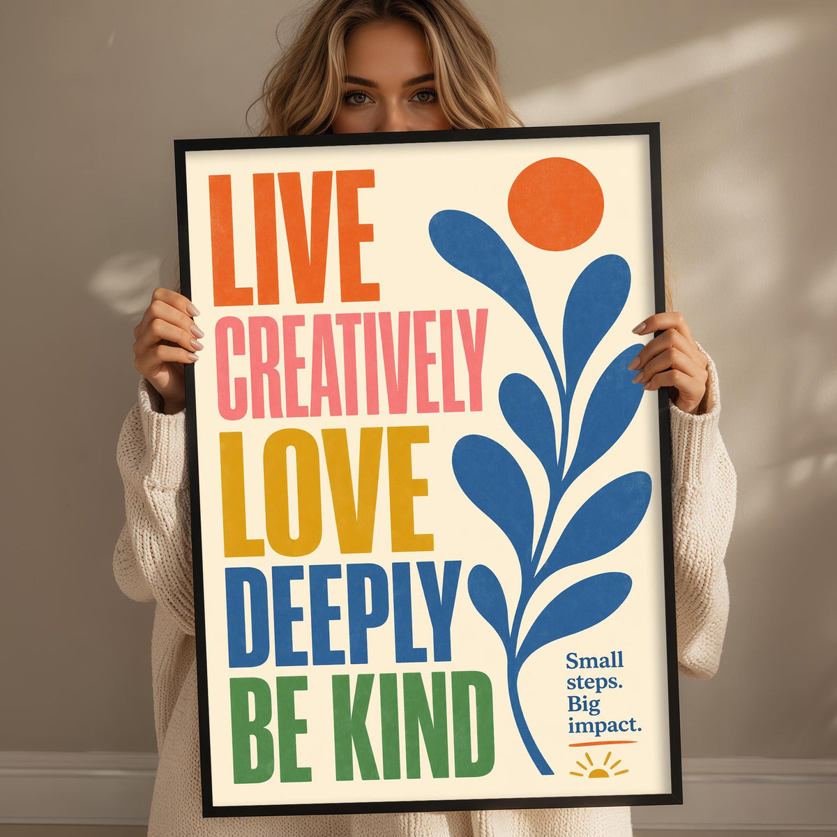 A woman holding a framed poster with the text "LIVE CREATIVELY LOVE DEEPLY BE KIND" and a sun graphic.