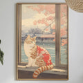 a framed painting of a cat sitting on a windowsill, with a view of a tree and a building outside. The painting is hanging on a wall above a dining table, which has various items on it, including a bottle, a bowl, and a vase. The table is set with plates, and there are some plants and other decorative items in the room.