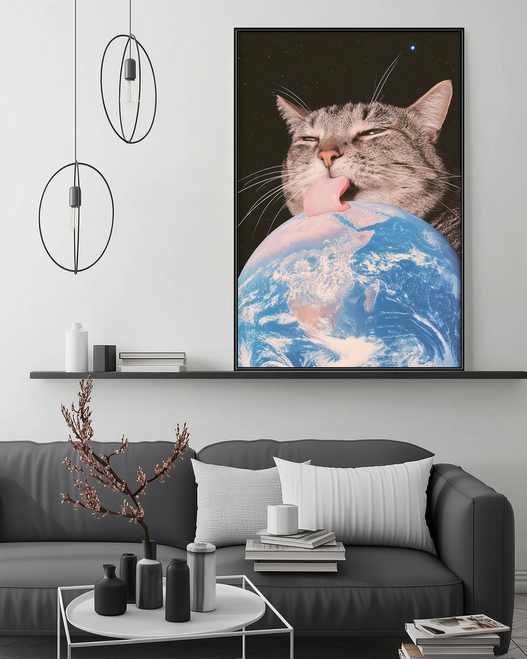 a large framed picture of a cat licking a globe, which appears to be a representation of the Earth. The cat is positioned on the globe, giving the impression that it is licking the Earth. The picture is displayed on a wall above a gray couch, and there are some decorative items, such as a potted plant and a vase, placed on the floor in front of the couch.