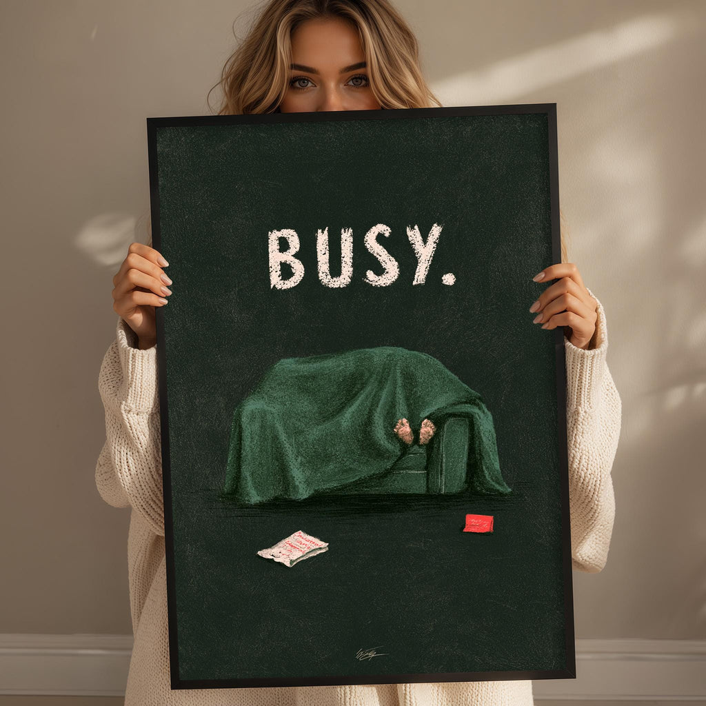 A woman holding a framed poster with an illustration of a person sitting on a couch under a blanket, with a red object on the floor next to the couch.