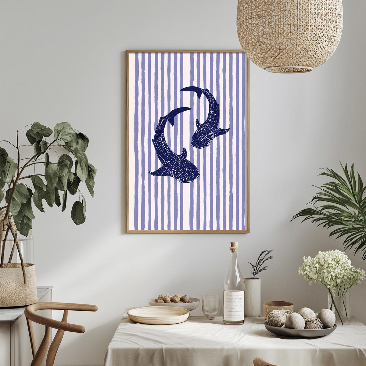a dining table with a white tablecloth, a bowl of fruit, a bottle, and a vase with flowers. Above the table, there is a framed picture of two blue sharks swimming against a striped background.