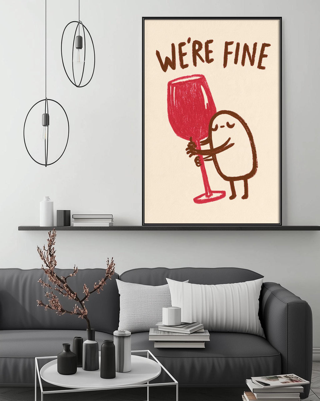 a cozy living room with a gray couch, a coffee table, and a framed poster on the wall. The poster depicts a whimsical illustration of a person holding a large red wine glass, accompanied by the text "WE'RE FINE".