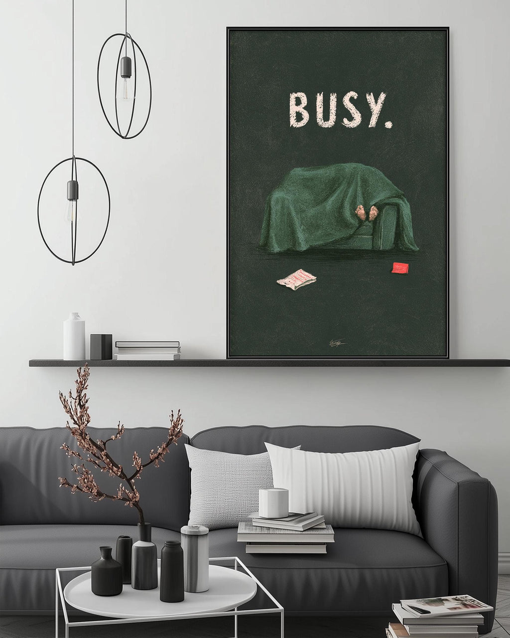 a modern living room with a gray sofa, a coffee table, and a large framed poster on the wall. The poster depicts a person sitting on a couch covered in a green blanket, with the word "BUSY" written above them.