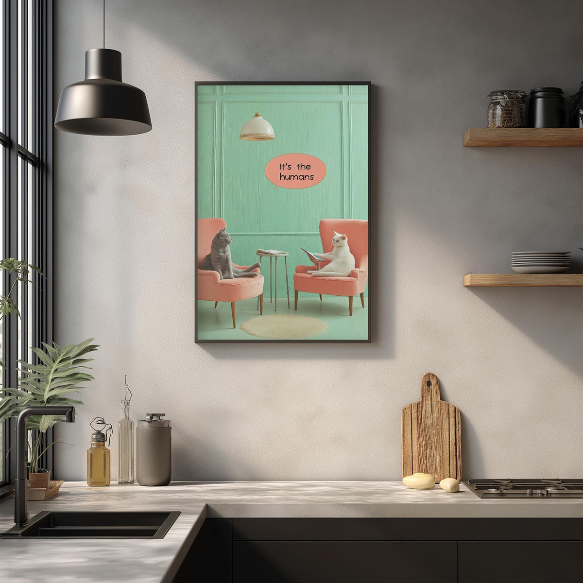 a kitchen scene with a framed poster of two cats sitting in chairs, one gray and one white, with the text "It's the humans" above them.