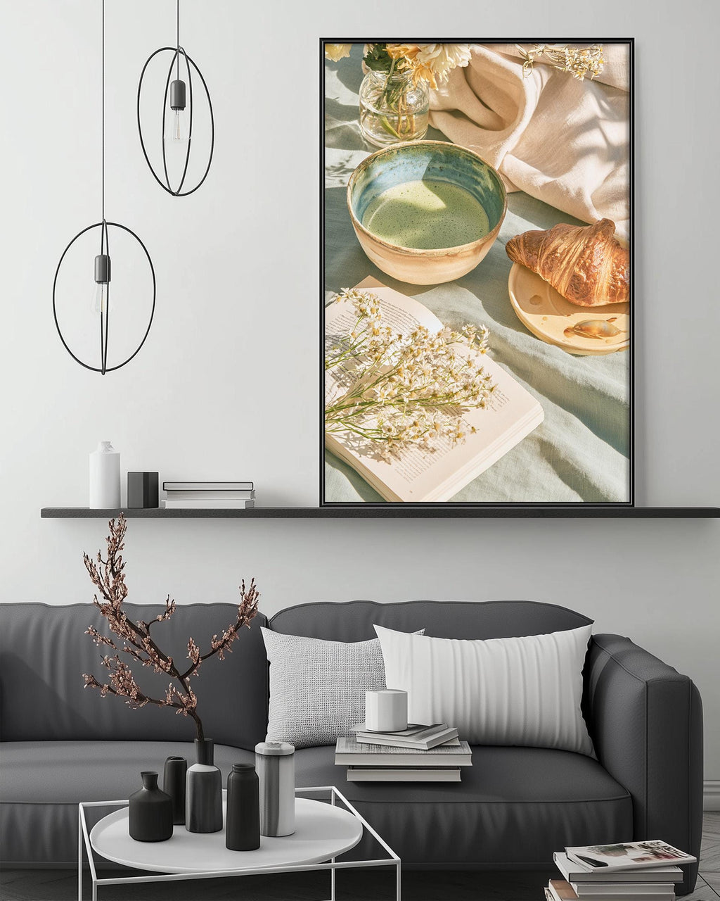 a cozy living room with a gray sofa, a coffee table, and a large framed picture of a table setting with a bowl, a plate of food, and a vase of flowers. The room has a minimalist and modern design, with pendant lights hanging above the sofa and a bookshelf below the picture.