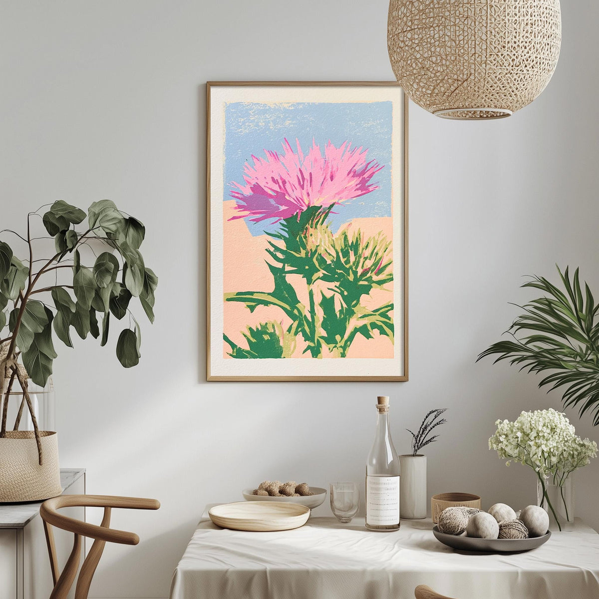 a dining table with a white tablecloth, a bowl of fruit, a bottle, and a vase with flowers. Above the table, there is a framed print of a pink flower on a green background.