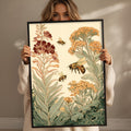A woman holds up a framed print of a floral and bee illustration, featuring a variety of flowers and bees.