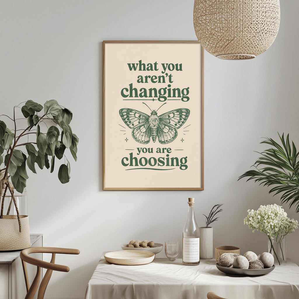 a framed poster with the text "what you aren't changing, you are choosing" and an illustration of a butterfly. The poster is hanging on a wall above a dining table with various items on it, including a bottle, a bowl, and a vase. The room also has potted plants and a hanging light fixture, creating a cozy and inviting atmosphere.
