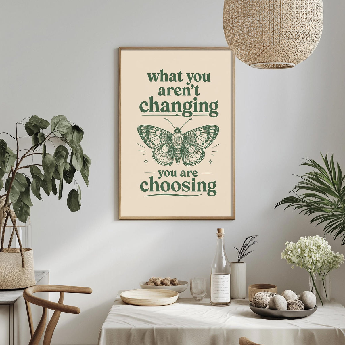 a framed poster with the text "what you aren't changing, you are choosing" and an illustration of a butterfly. The poster is hanging on a wall above a dining table with various items on it, including a bottle, a bowl, and a vase. The room also has potted plants and a hanging light fixture, creating a cozy and inviting atmosphere.