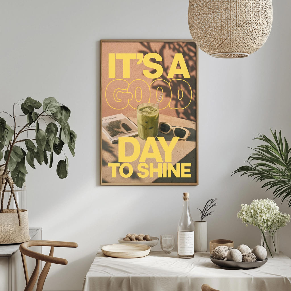 a dining table with a white tablecloth, a bowl of fruit, a bottle, and a vase with flowers. Above the table, there is a framed poster with the text "IT'S A GOOD DAY TO SHINE" in yellow and orange colors.