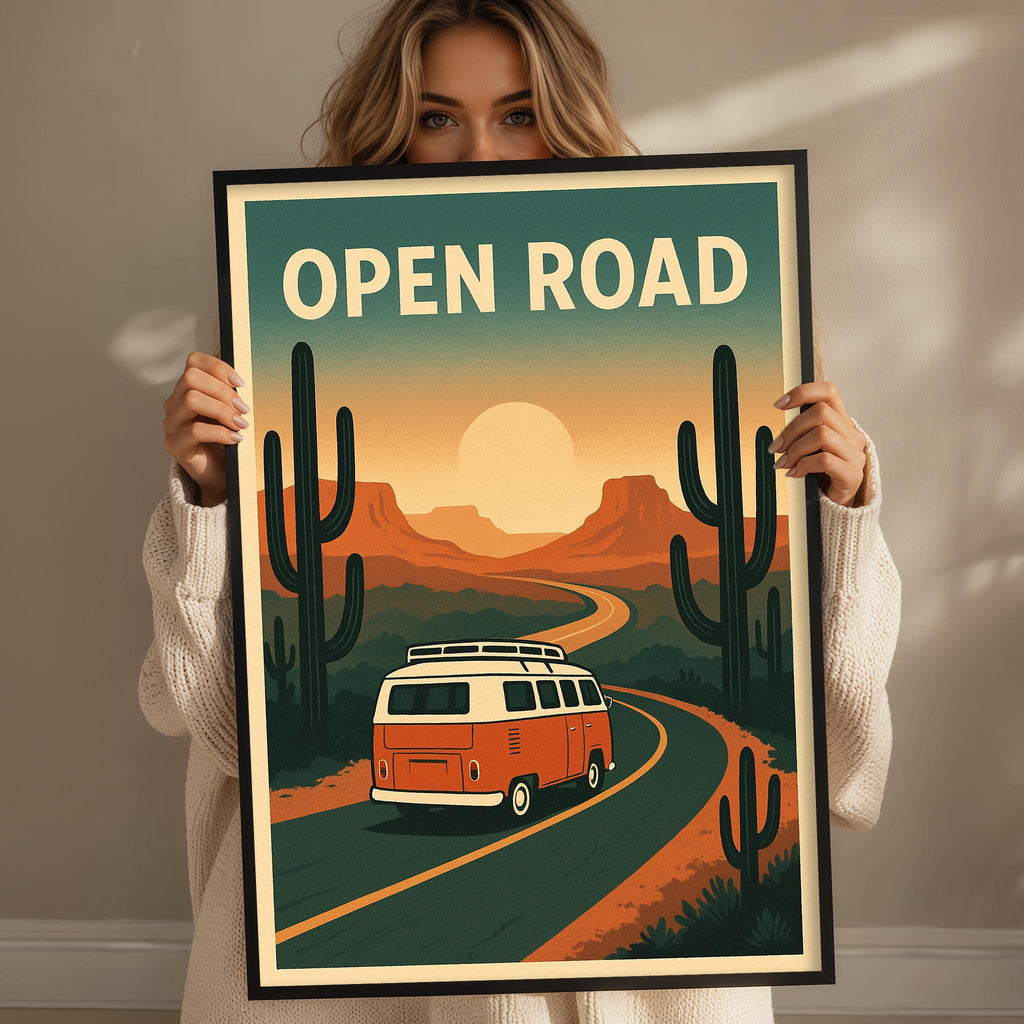 A woman holding a framed poster with a vintage-style illustration of a van driving on a road through a desert landscape with cacti and mountains in the background.