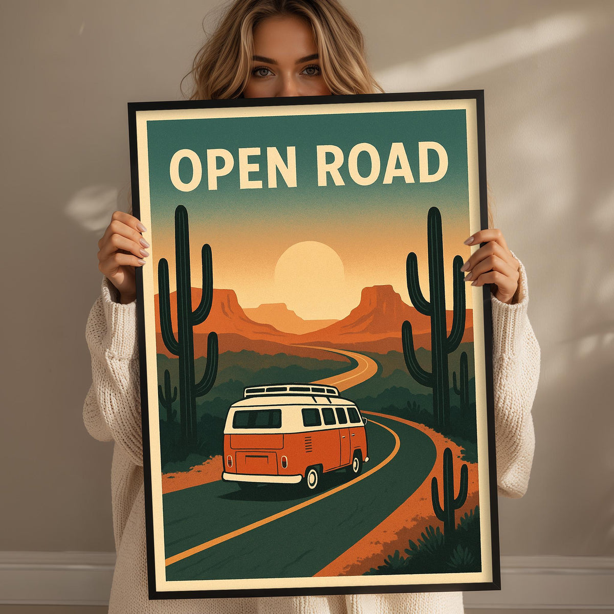 A woman holding a framed poster with a vintage-style illustration of a van driving on a road through a desert landscape with cacti and mountains in the background.