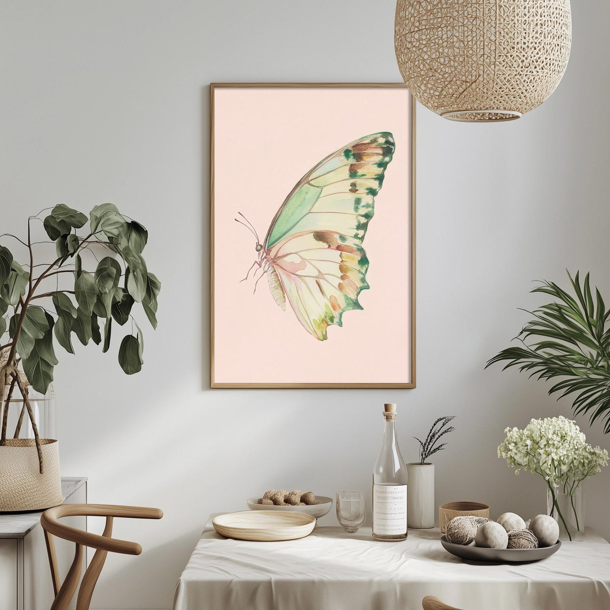 a dining table with a white tablecloth, a bowl of fruit, a bottle, and a vase with flowers. Above the table, there is a framed picture of a butterfly on a pink background.