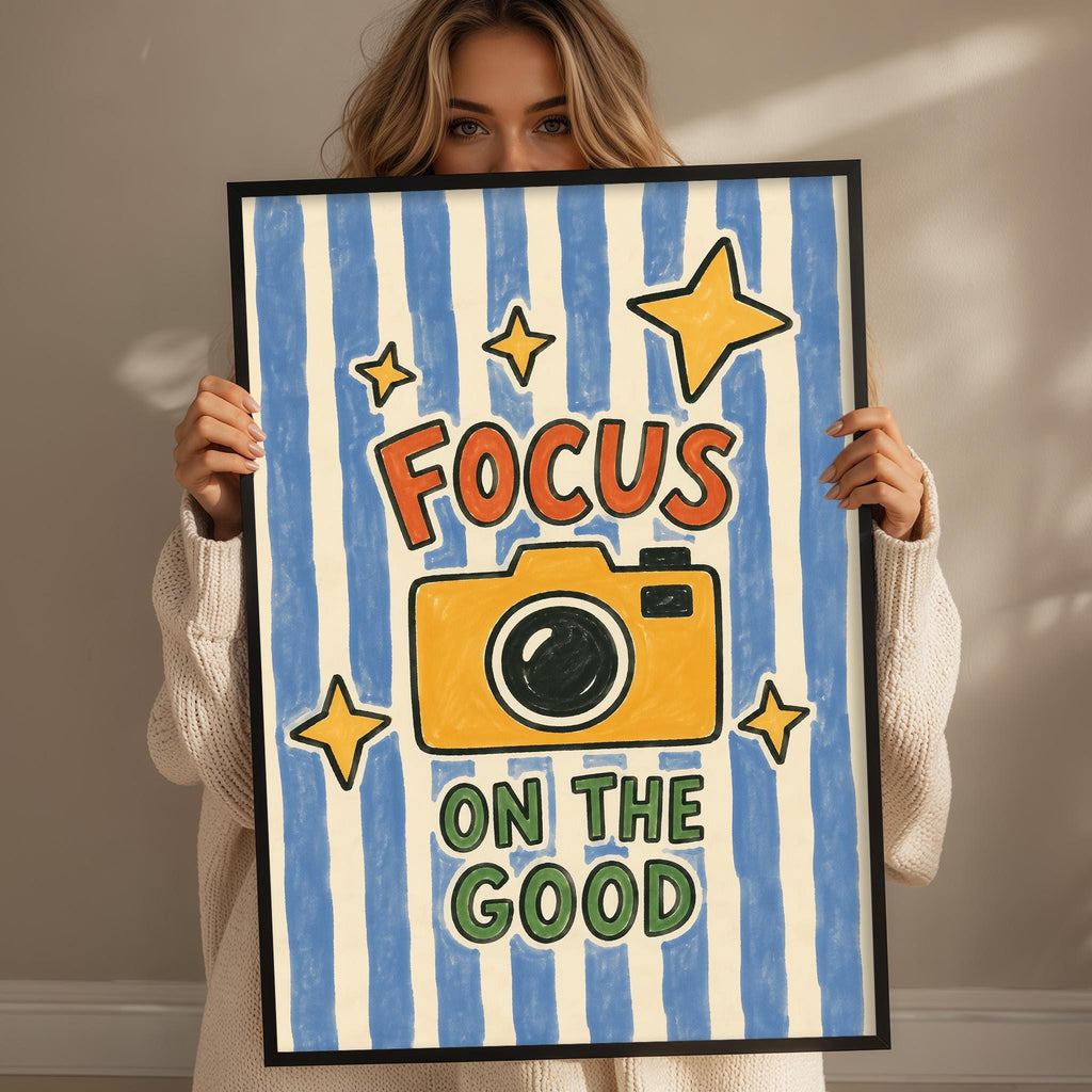 A woman holding a framed poster with the text "Focus on the good" and an illustration of a camera.