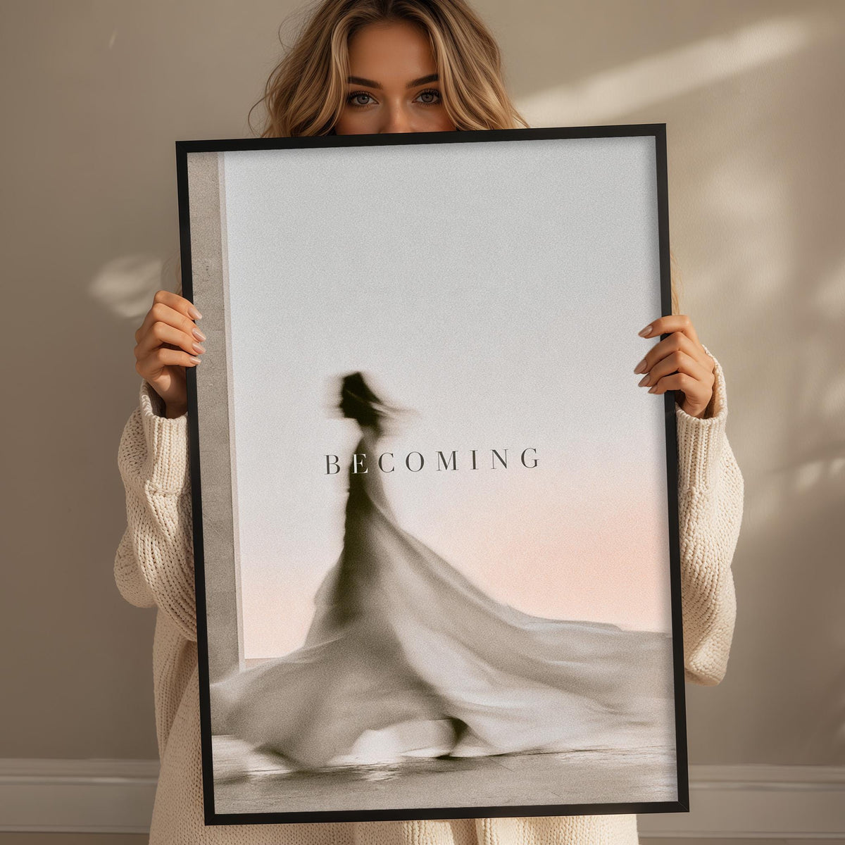 A woman holding a framed poster with a blurred silhouette of a person in a flowing dress and the word "BECOMING" written on it.