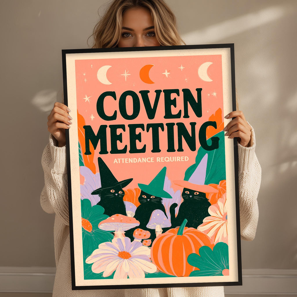 A woman is holding up a framed poster with a Halloween-themed illustration of cats wearing witch hats and surrounded by flowers and mushrooms.