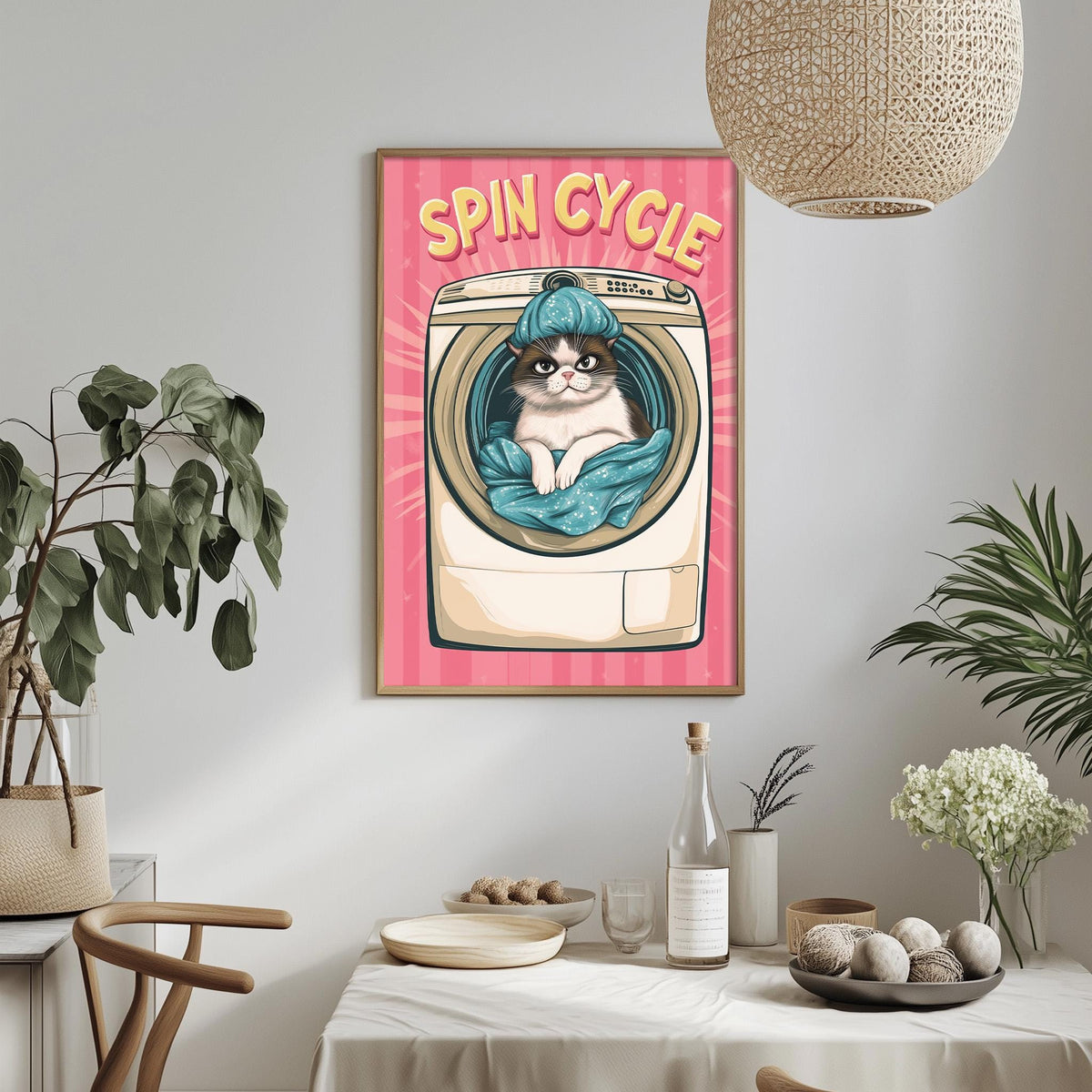 a framed poster of a cat sitting inside a washing machine, with the text "SPIN CYCLE" written above it. The poster is displayed on a wall above a dining table with various items on it, including a bottle, a bowl, and a vase.