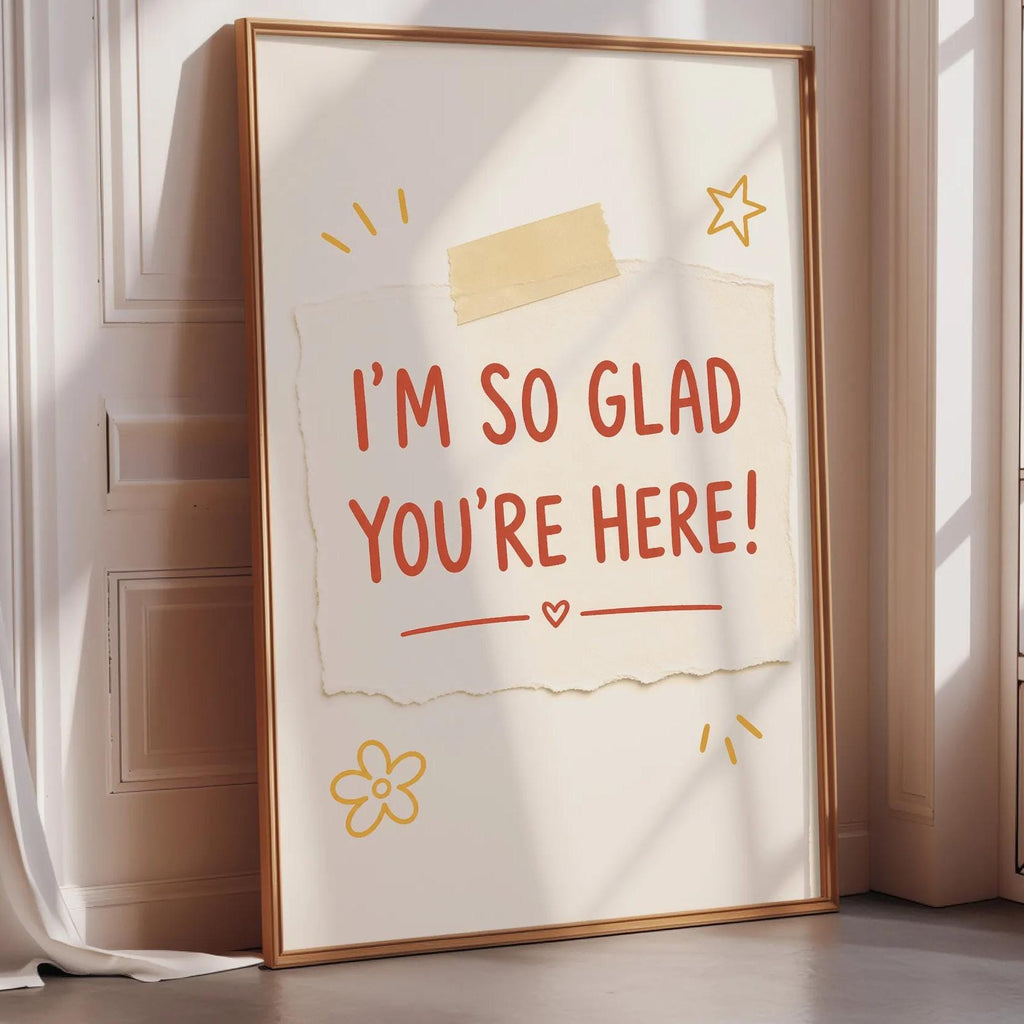 A framed poster with the text "I'm so Glad You're Here!" displayed on it, along with some decorative elements such as a flower and a star.