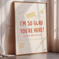 A framed poster with the text "I'm so Glad You're Here!" displayed on it, along with some decorative elements such as a flower and a star.