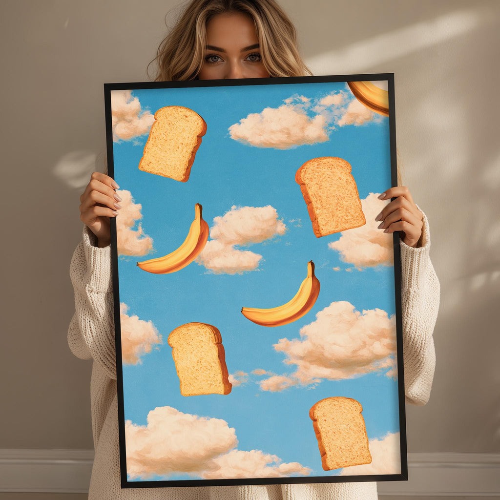 A woman holds up a framed print with a whimsical illustration of bread and bananas floating in a blue sky with clouds.