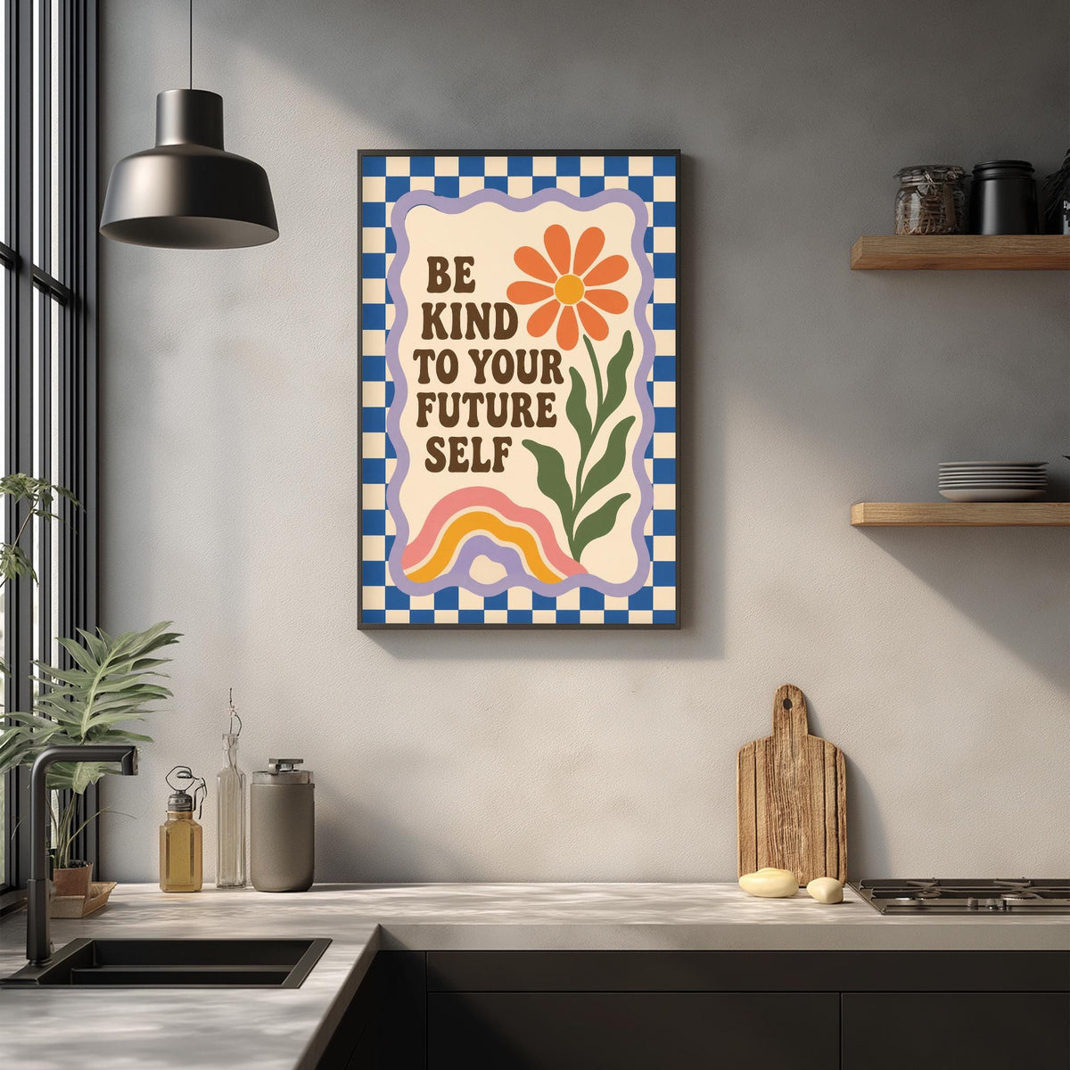 a kitchen counter with a framed poster that reads "Be kind to your future self" above it. The poster has a colorful design with a flower and a rainbow. There are also various kitchen items on the counter, such as a cutting board, a knife, and a bottle.