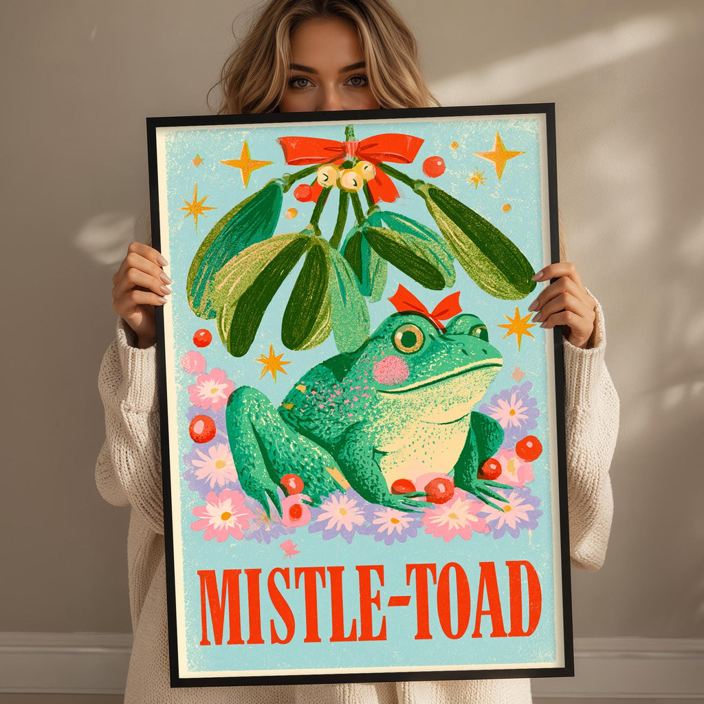 A woman is holding up a framed poster with a green frog and mistletoe illustration.