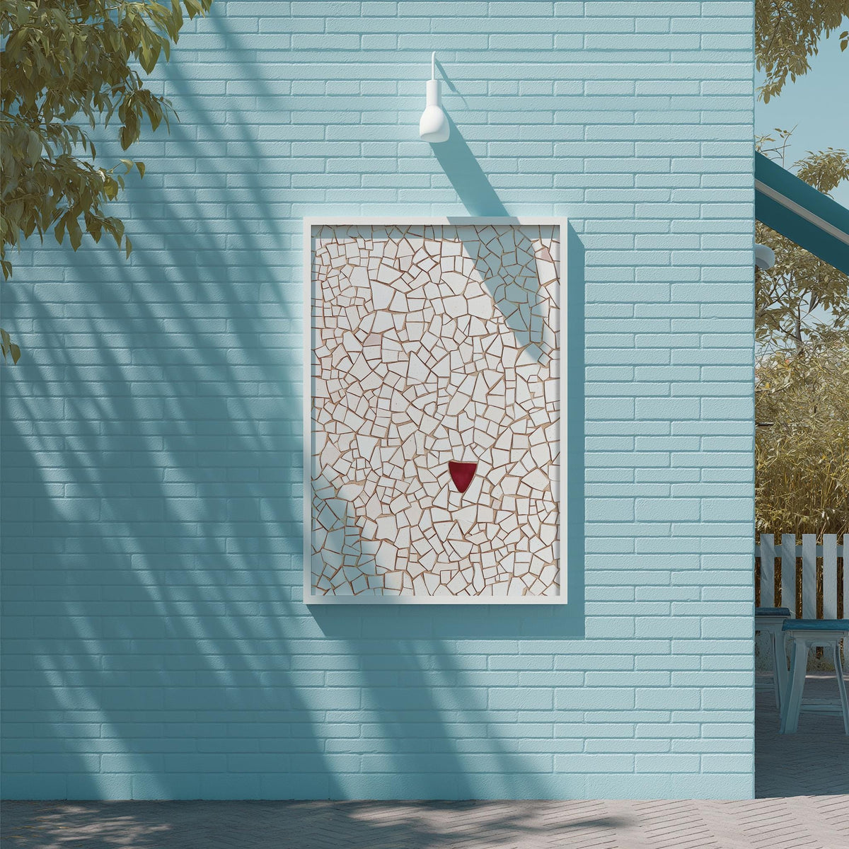 a blue brick wall with a white square frame containing a mosaic design. The mosaic appears to be made of small tiles or stones, creating a visually striking pattern. The wall is situated next to a tree, and there is a white light fixture mounted on the wall above the frame.