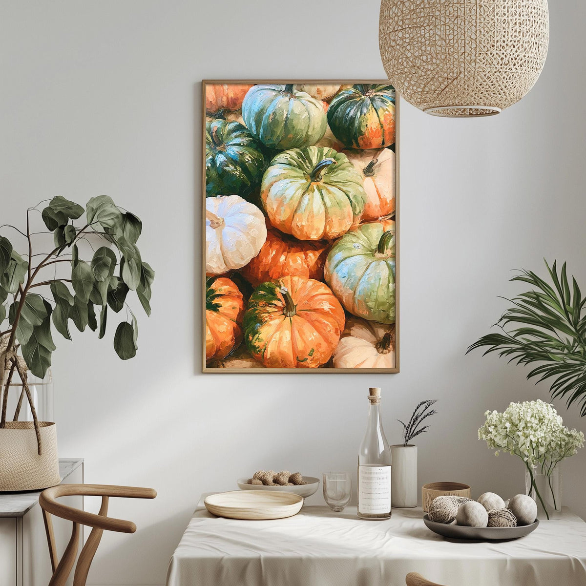 a dining table with a white tablecloth, a bowl of fruit, a bottle, and a vase with flowers. Above the table, there is a framed painting of pumpkins hanging on the wall.