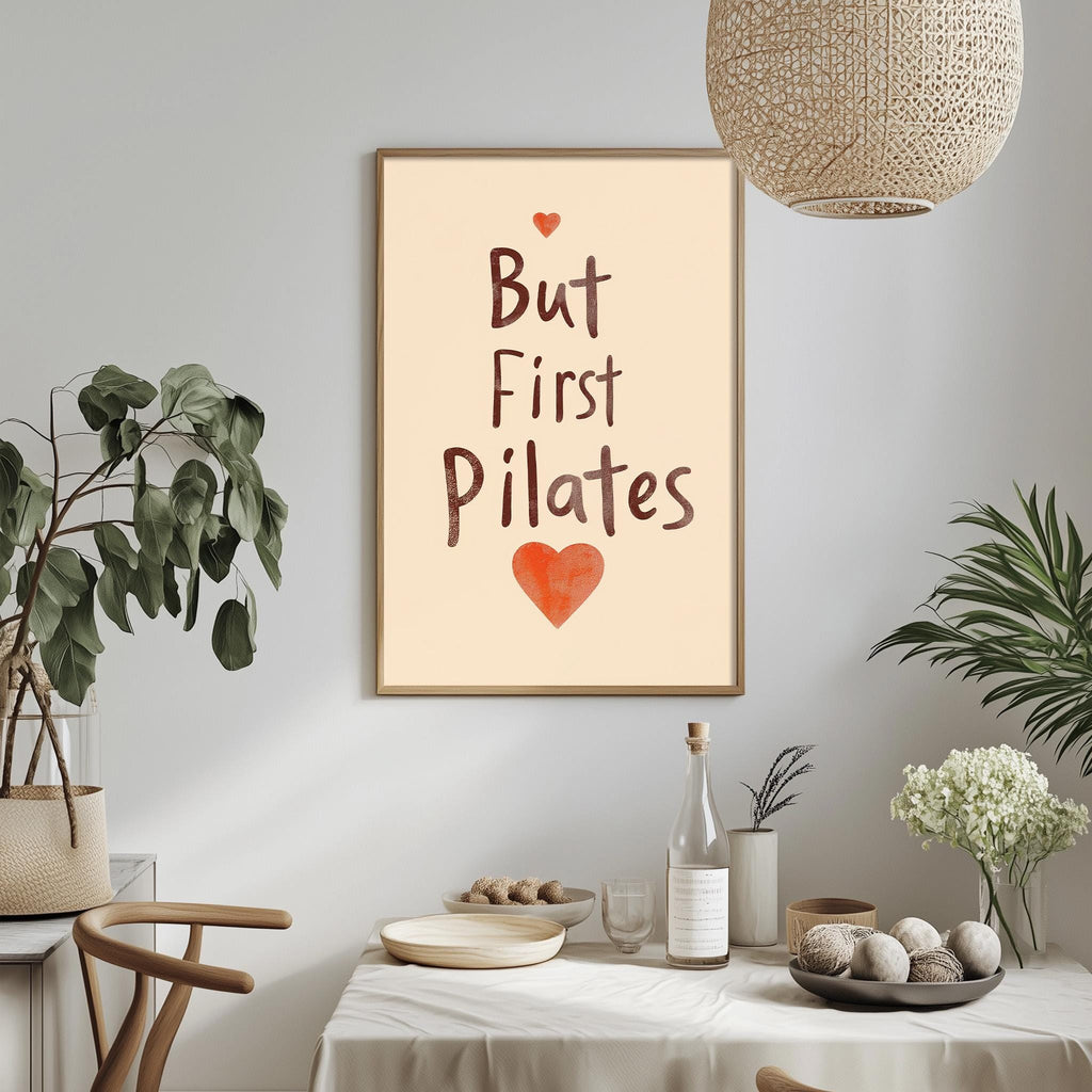 a framed poster with the text "But First Pilates" and an orange heart symbol, hanging on a wall above a dining table with various items on it.