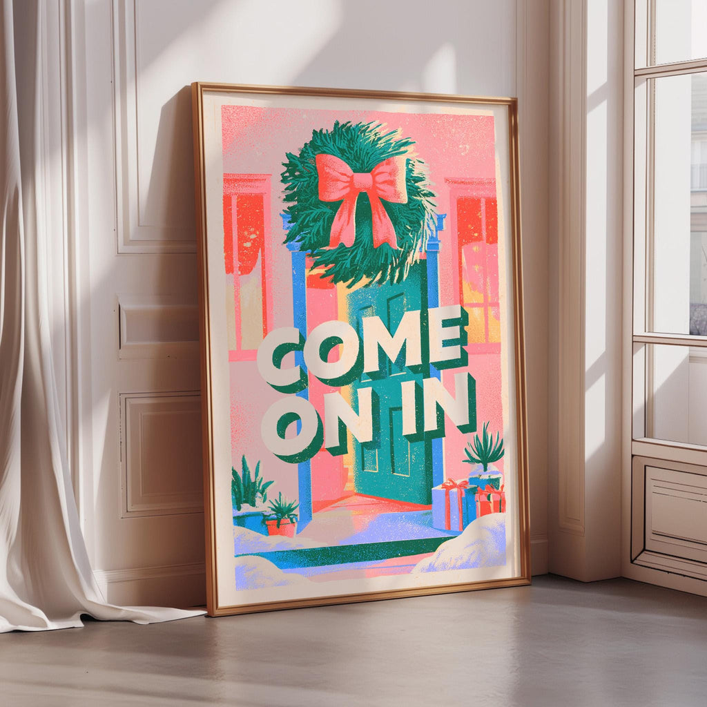 A framed poster with a wreath and the text "COME ON IN" displayed on it, placed on a wooden floor in a room with a window.