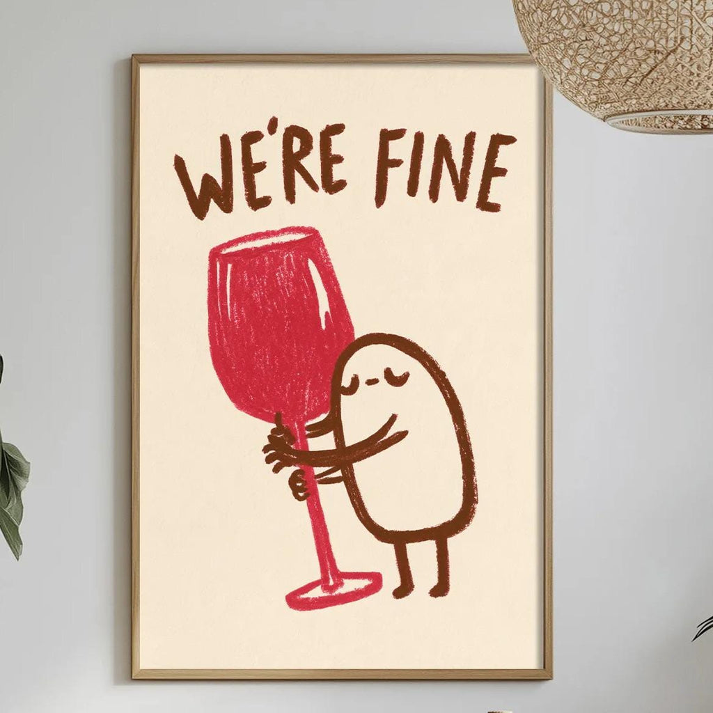 a framed poster with a cartoon character holding a wine glass, accompanied by the text "WE'RE FINE". The poster is displayed on a wall above a dining table with various items on it, including a bottle, a bowl, and a vase.