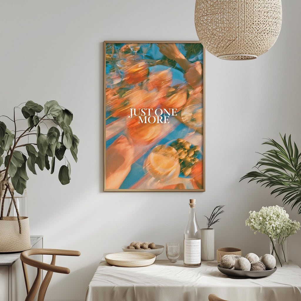 a dining table with a white tablecloth, a bowl of fruit, a bottle, and a vase with flowers. Above the table, there is a framed poster with the text "JUSTONE MORE" in the center.