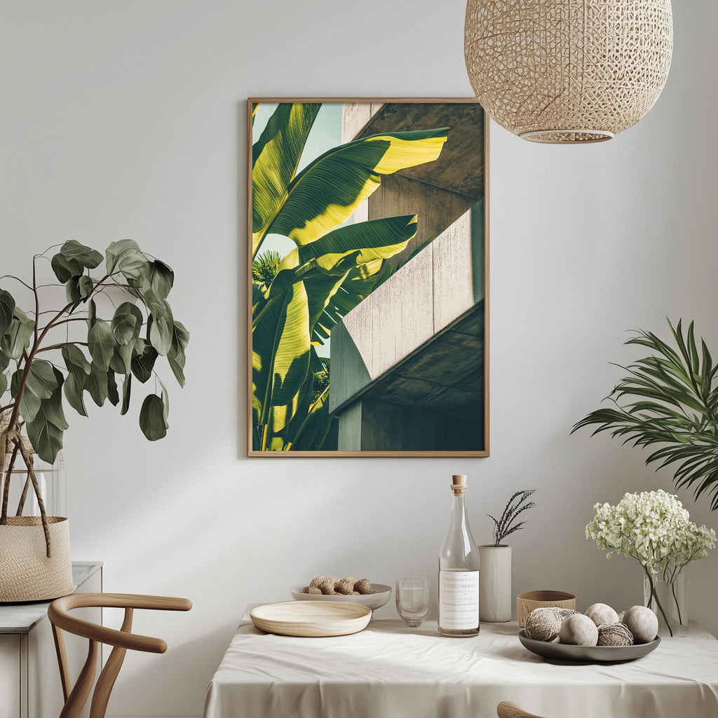 a dining table with a white tablecloth, a bowl of fruit, a bottle, and a vase with flowers. A framed picture of a banana tree hangs on the wall above the table.