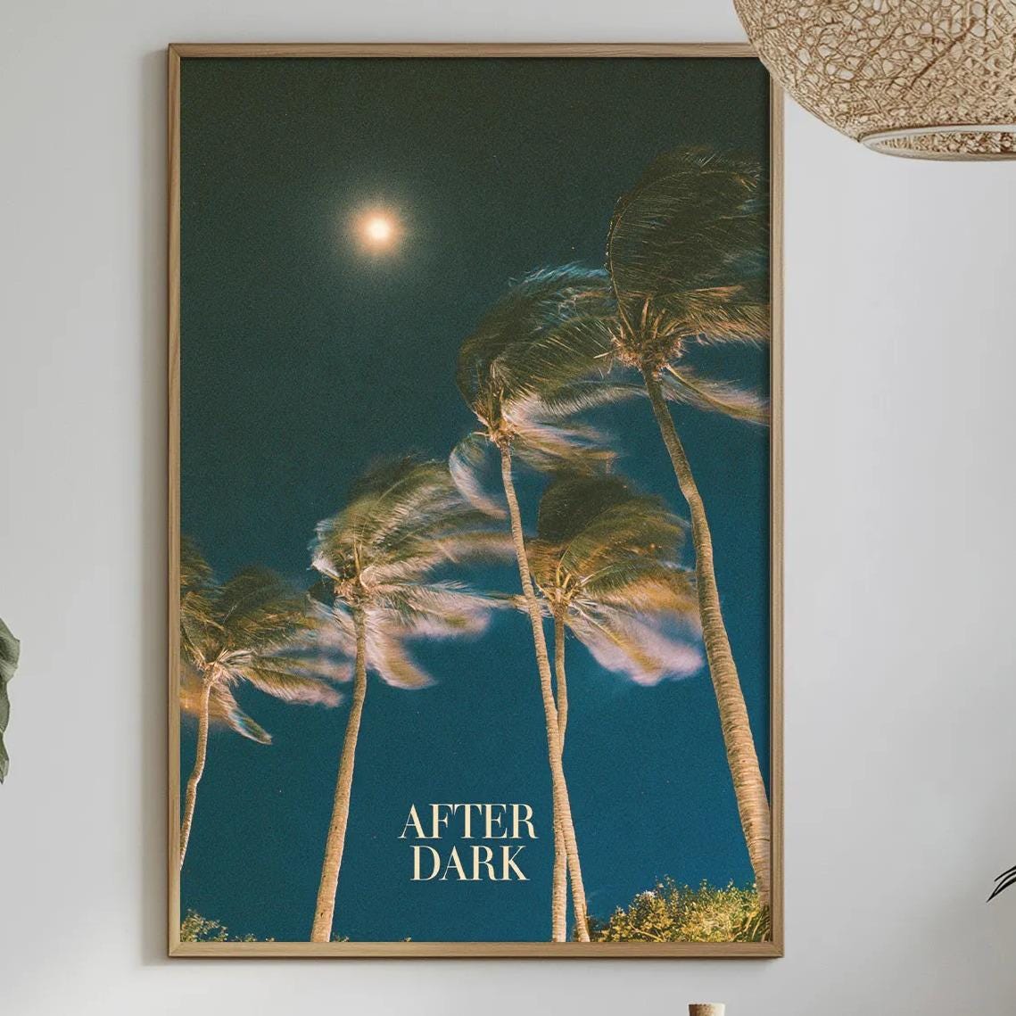 a dining table with a bowl of fruit, a bottle, and a vase of flowers, along with a framed poster of palm trees hanging on the wall above the table.