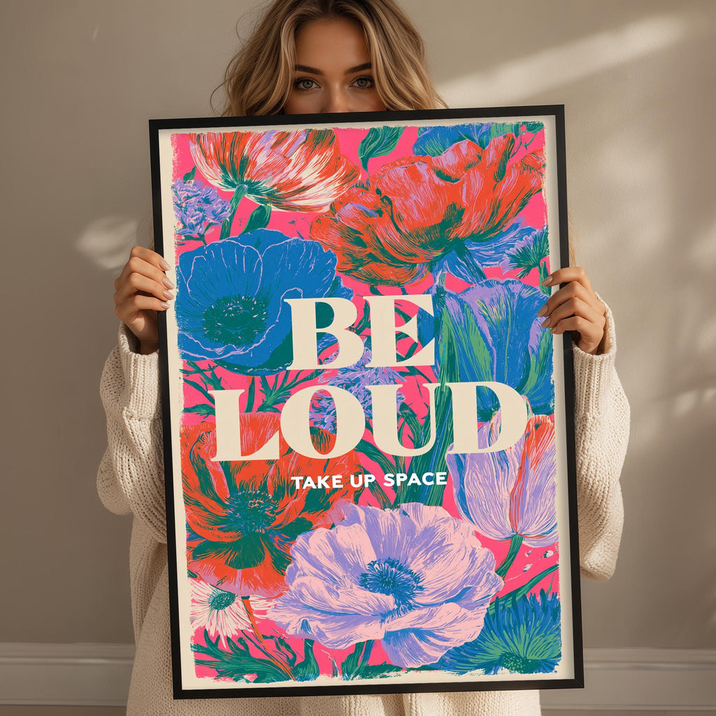A woman holding a framed poster with a colorful floral design and the text "BE LOUD TAKE UP SPACE".