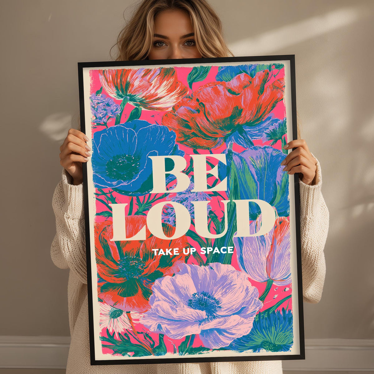 A woman holding a framed poster with a colorful floral design and the text "BE LOUD TAKE UP SPACE".