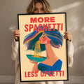 a woman holding a framed poster with a colorful illustration of a woman eating spaghetti. The poster has the text "MORE SPAGHETTI LESS UPSETTI" written on it.