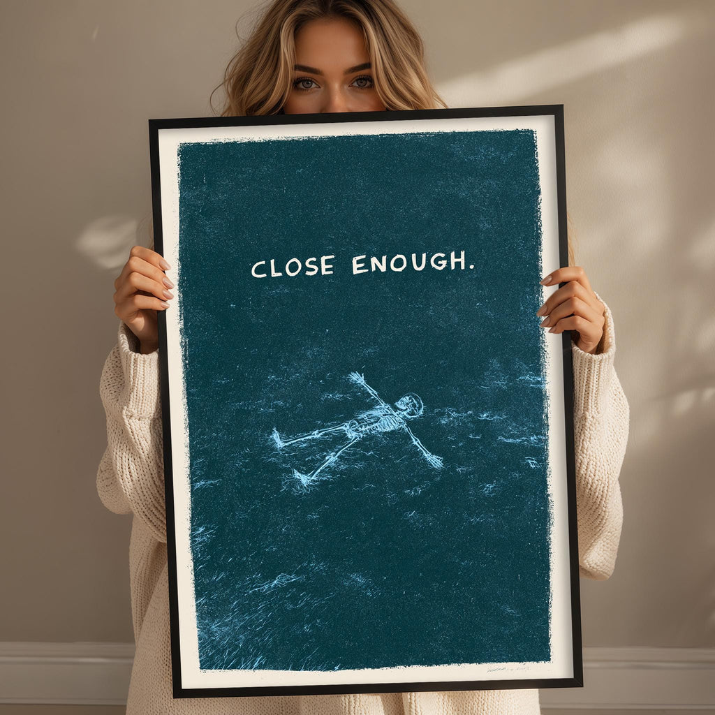 A woman is holding up a framed poster with the text "CLOSE ENOUGH" on it.