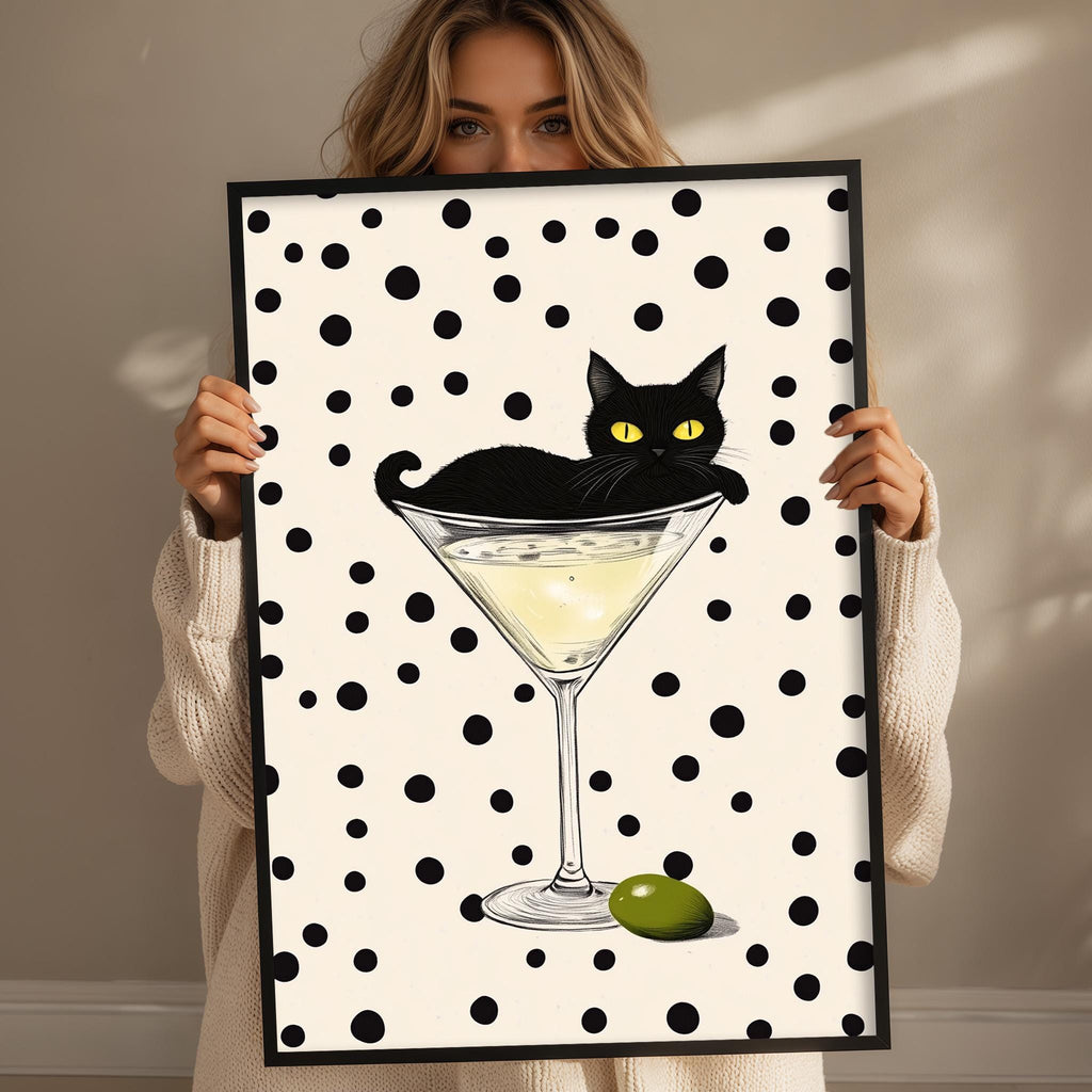 A woman holds up a framed print with a black cat sitting in a martini glass, which is filled with a yellow liquid and has an olive on the rim.