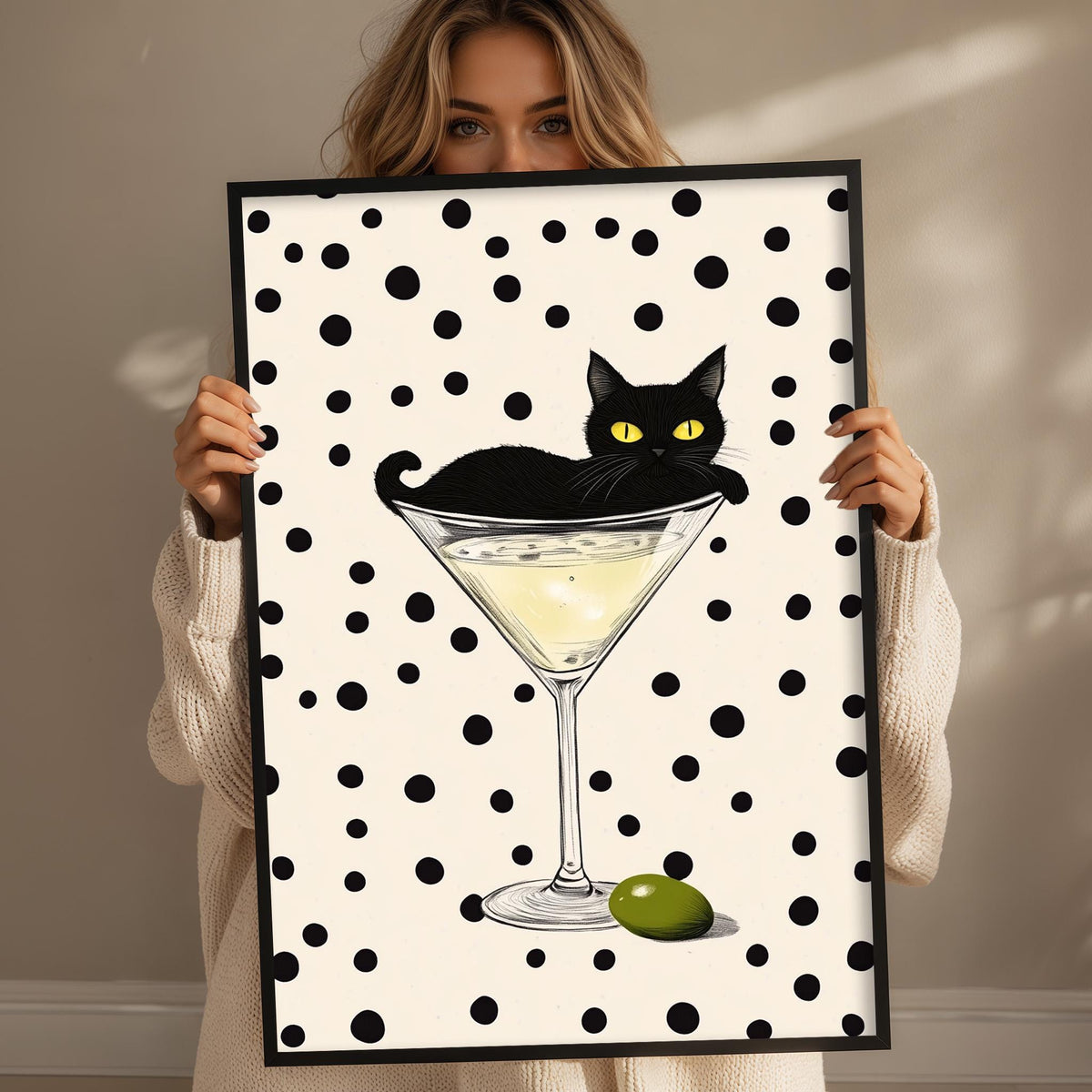 A woman holds up a framed print with a black cat sitting in a martini glass, which is filled with a yellow liquid and has an olive on the rim.