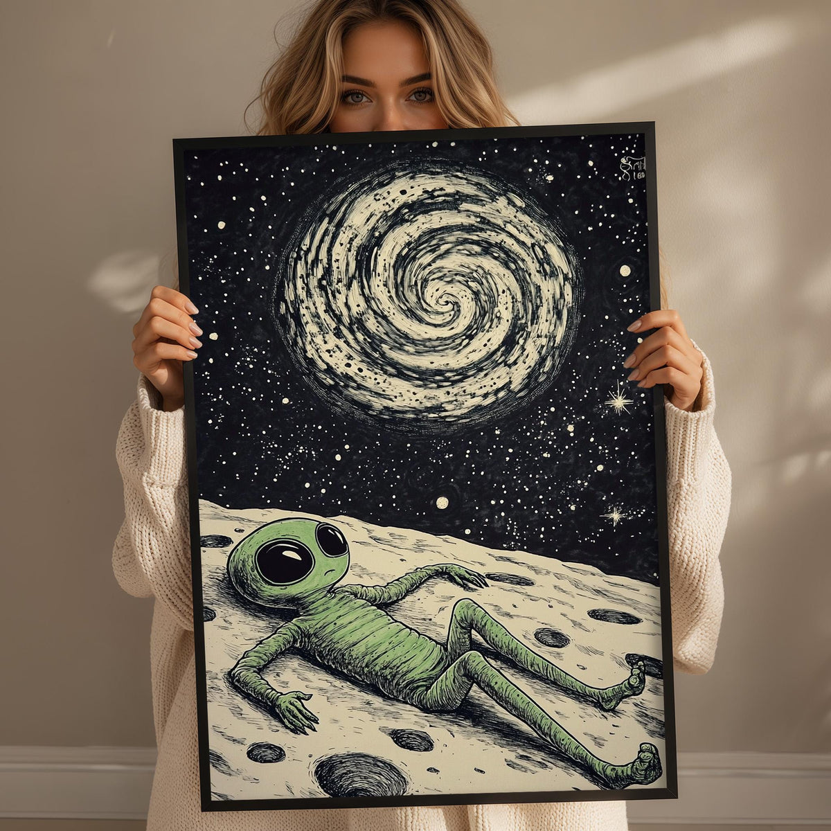A woman holds up a framed artwork depicting an alien lying on a rocky surface, gazing up at a spiral galaxy in the night sky.