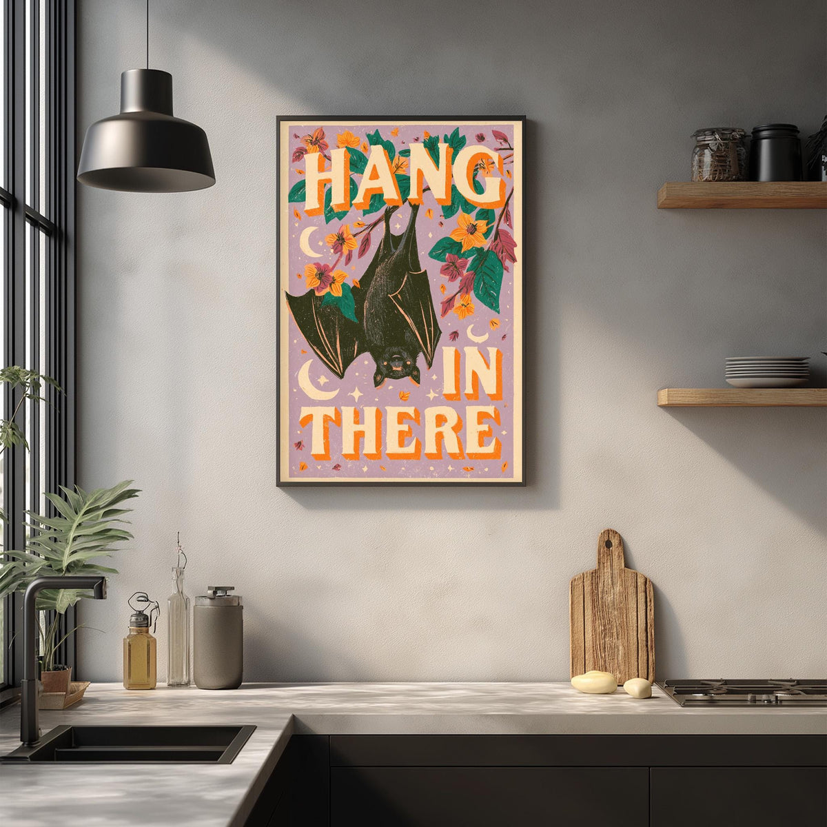 a kitchen counter with a framed poster hanging on the wall above it. The poster has a bat illustration and the text "HANG IN THERE" written in large letters.