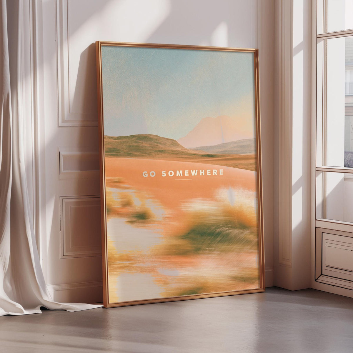 A framed poster with a desert landscape and the text "Go somewhere" is displayed on a wall in a room with sunlight streaming in from a window.
