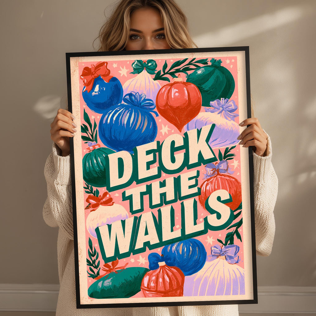 A woman is holding up a framed poster with a colorful design featuring various Christmas-themed elements such as ornaments, ribbons, and a text that reads "Deck the Walls".